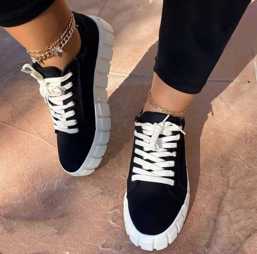 Chaussures de sport noires avec semelles épaisses blanches, lacets blancs, portées avec des bracelets de cheville dorés sur un sol en pierre.