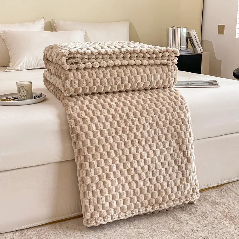 Couverture en tricot beige texturé sur un lit blanc dans une chambre moderne, avec oreillers blancs et une bougie parfumée sur la table de chevet.