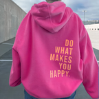 Personne portant un sweat à capuche rose vif avec le texte "Do What Makes You Happy" en jaune, debout dans un parking extérieur.