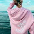 Personne en sweat à capuche rose avec message "Do What Makes You Happy" face à la mer, ciel bleu. Mode décontractée, inspiration, bien-être.