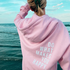 Personne en sweat à capuche rose avec message "Do What Makes You Happy" face à la mer, ciel bleu. Mode décontractée, inspiration, bien-être.