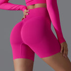 Femme en tenue de sport rose vif, portant un short moulant taille haute et un haut à manches longues, posant de profil sur fond gris.