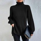 Pull noir à col roulé en maille épaisse, porté avec un sac à main matelassé noir à chaîne dorée, sur fond de mur texturé blanc.