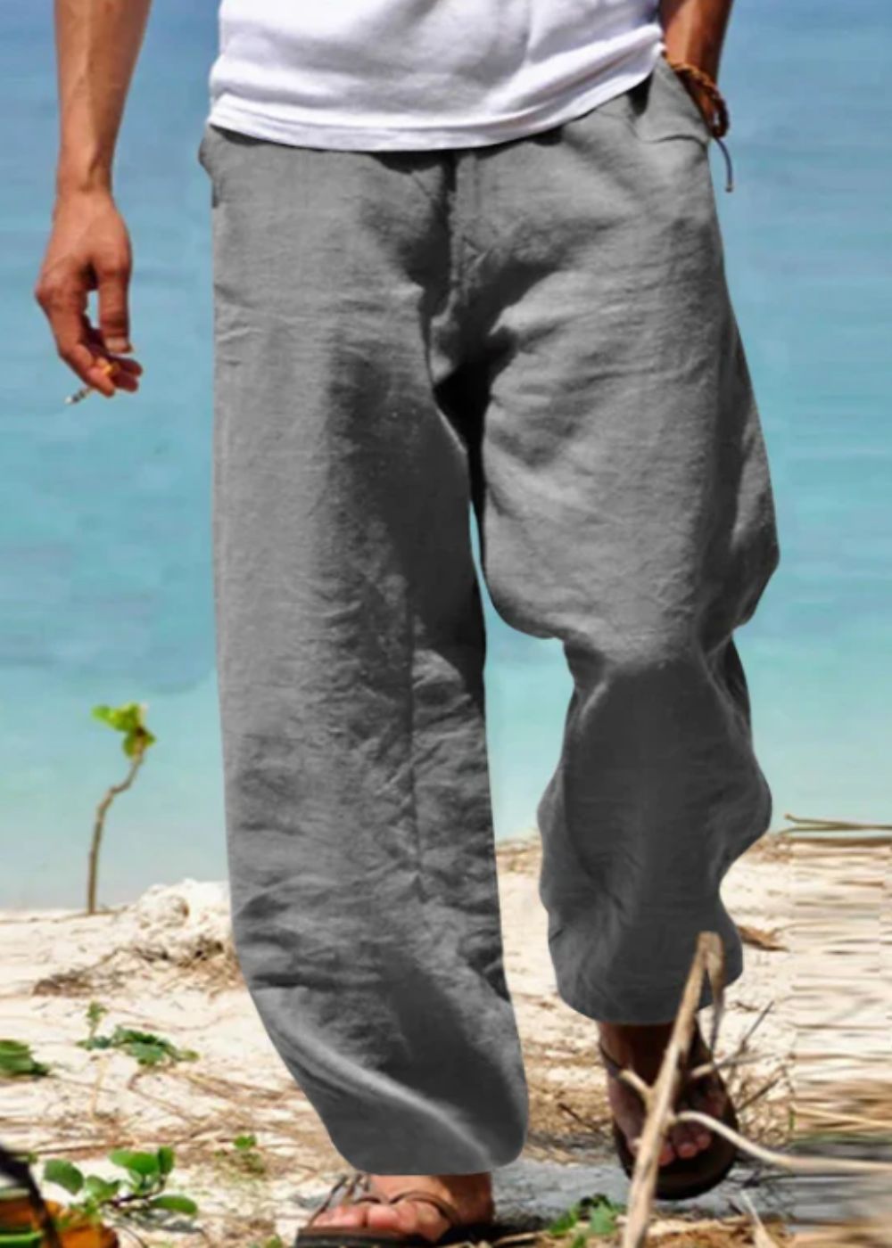 Homme portant un pantalon en lin gris sur une plage, pieds nus dans le sable, avec vue sur la mer turquoise. Mode estivale décontractée.
