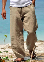 Homme portant un pantalon en lin beige sur une plage, pieds nus, avec vue sur la mer turquoise. Mode estivale, détente, vêtements légers.