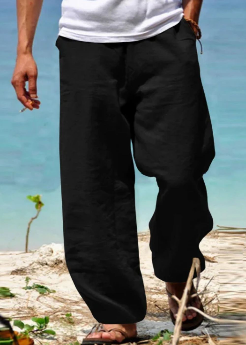 Homme portant un pantalon ample noir en lin sur une plage, pieds nus dans le sable, mer turquoise en arrière-plan. Mode estivale décontractée.