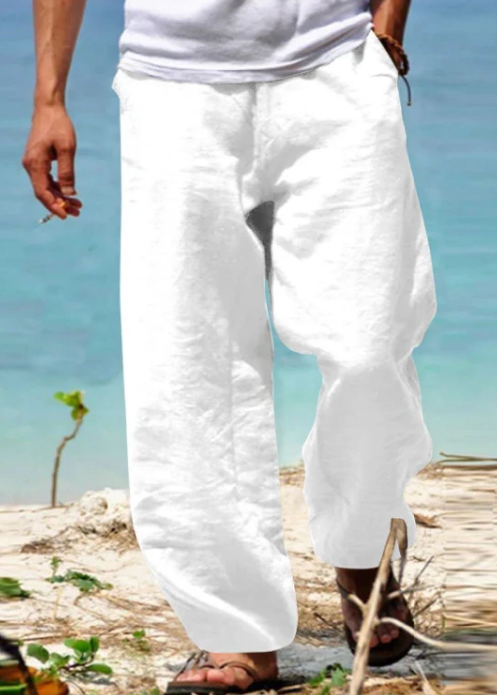 Homme portant un pantalon blanc en lin sur une plage, pieds nus dans le sable, mer turquoise en arrière-plan. Mode estivale, détente bord de mer.