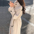 Femme portant un manteau beige élégant, ceinturé, manches longues, sur fond de rue ensoleillée. Mode automne, style urbain chic.