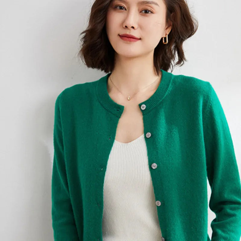 Femme portant un cardigan vert en laine, sur un débardeur blanc, posant contre un mur blanc. Mode féminine, style décontracté, vêtements d'hiver.