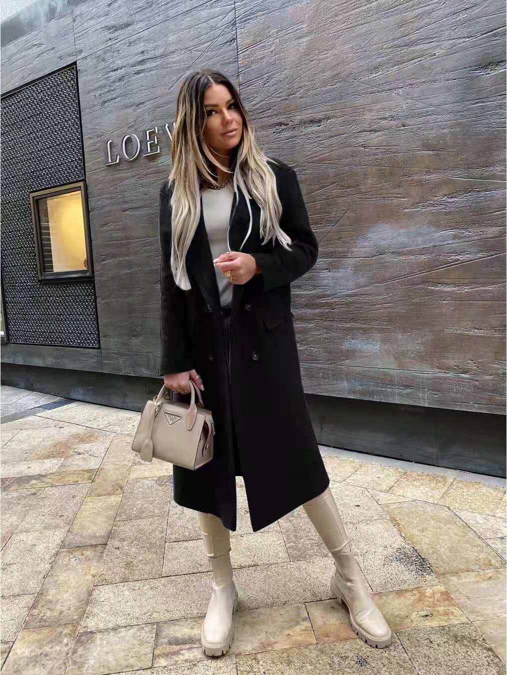 Femme élégante en manteau noir, portant un sac Prada beige, posant devant un mur avec le logo Loewe. Mode automne, style urbain chic.