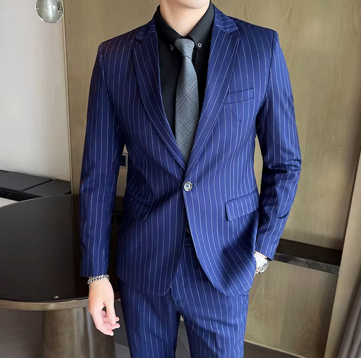 Homme en costume bleu à rayures, chemise noire et cravate grise, posant dans un intérieur moderne. Mode masculine élégante et professionnelle.