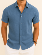 Homme portant une chemise bleue à manches courtes, élégante et décontractée, avec un pantalon blanc. Mode masculine estivale, style moderne.