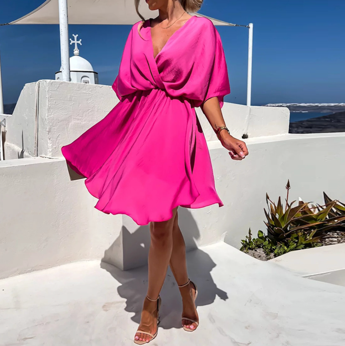 Robe rose vif élégante portée par une femme sur une terrasse blanche en bord de mer, ciel bleu en arrière-plan, style estival chic.