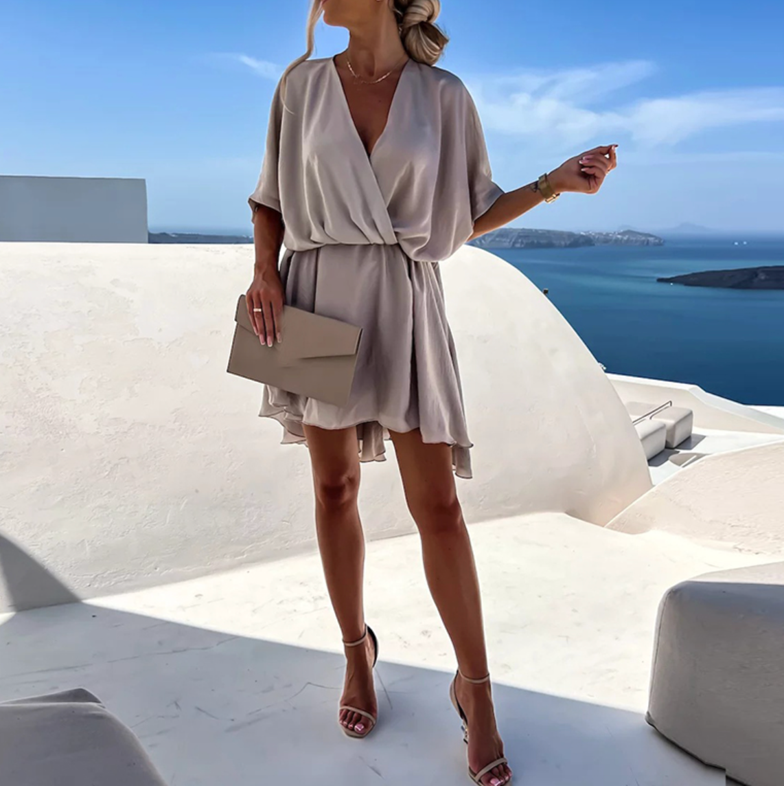 Femme en robe beige élégante sur terrasse avec vue sur mer, tenant une pochette. Mode estivale, style chic, paysage côtier en arrière-plan.