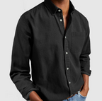 Homme portant une chemise noire en denim, manches longues, avec poches, sur fond blanc. Mode masculine, style décontracté, tendance actuelle.