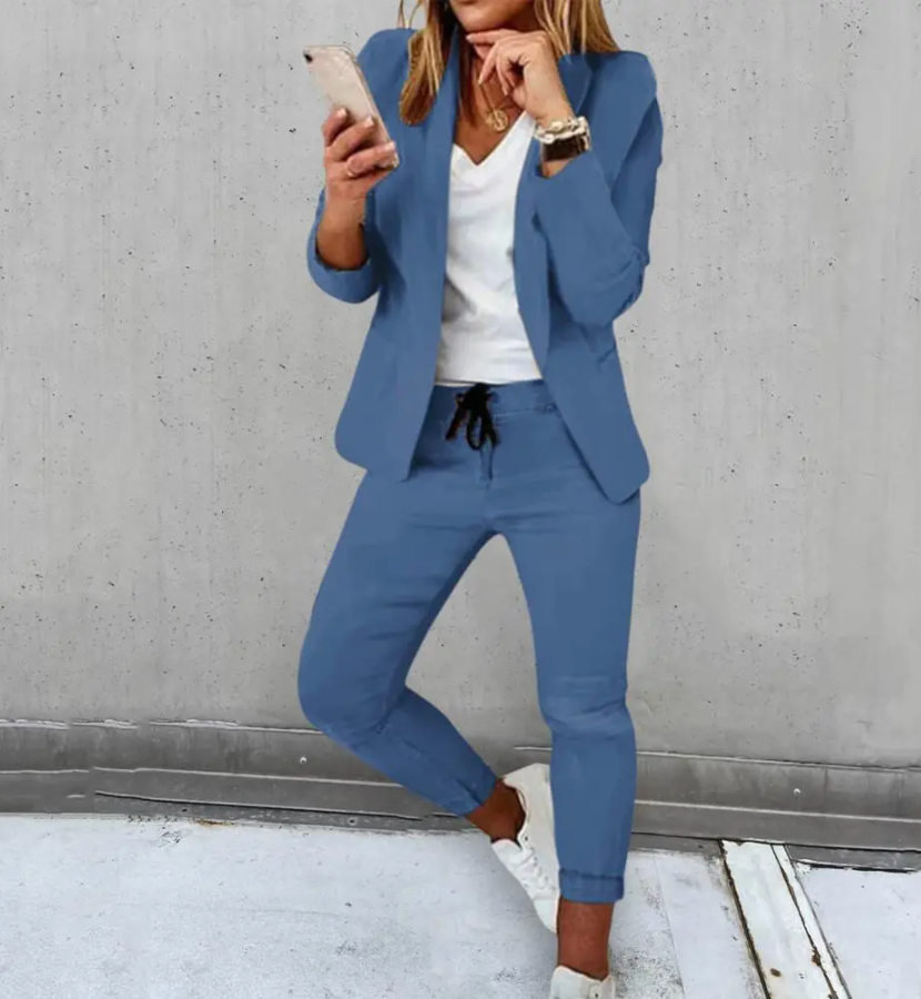 Femme en costume bleu décontracté, tenant un smartphone, portant des baskets blanches, posant contre un mur gris. Mode féminine, tenue élégante.