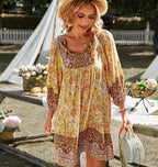 Femme en robe bohème jaune à motifs floraux, chapeau de paille, tenant un sac en osier, debout près d'une table en bois dans un jardin ensoleillé.