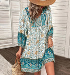Robe bohème fleurie bleue et blanche, manches longues, portée par une femme avec chapeau de paille, tenant un sac en osier. Style estival chic.