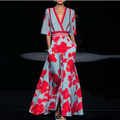Robe longue élégante à motifs floraux rouges sur fond bleu, manches évasées, décolleté en V, portée par un mannequin sur un podium de mode.