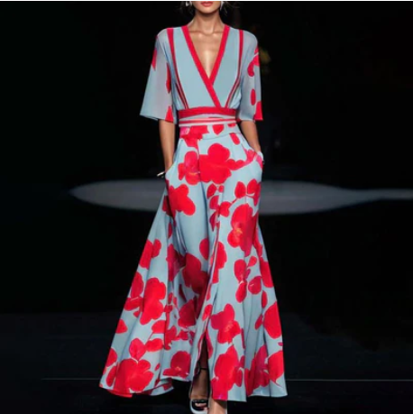 Robe longue élégante à motifs floraux rouges sur fond bleu, manches évasées, décolleté en V, portée par un mannequin sur un podium de mode.