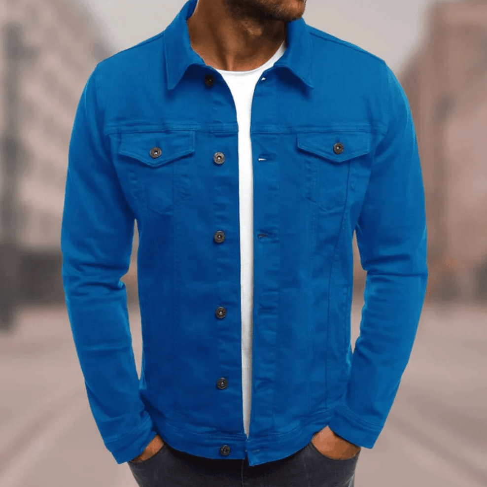 Homme portant une veste en jean bleu vif, chemise blanche en dessous, debout dans une rue floue. Mode masculine, style urbain, tendance vestimentaire.