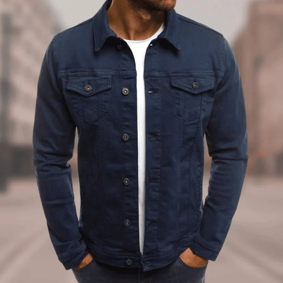 Homme portant une veste en jean bleu foncé, chemise blanche en dessous, debout dans une rue floue. Mode masculine, style décontracté, tendance urbaine.
