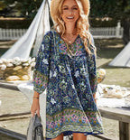 Robe bohème chic à motifs floraux bleus et verts, femme souriante avec chapeau de paille, ambiance estivale en plein air, style décontracté élégant.