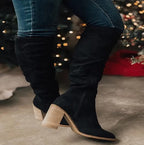 Bottes noires en daim à talons hauts, portées avec un jean, devant un sapin de Noël illuminé. Mode hiver élégante et tendance.