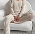 Femme assise sur un canapé blanc, portant un ensemble en tricot beige côtelé. Mode automne-hiver, confort et élégance, tenue décontractée.
