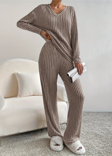 Femme en ensemble lounge côtelé marron, tenant une pochette argentée, debout sur un tapis gris, devant un fauteuil blanc. Mode détente élégante.