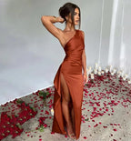 Femme en robe longue satinée orange, fendue, posant sur un sol couvert de pétales de rose rouges et bougies blanches, ambiance élégante et romantique.