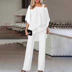 Femme en tenue élégante blanche, blouse asymétrique et pantalon large, tenant une pochette noire, dans un intérieur moderne et épuré.