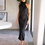Robe longue noire élégante sans manches, col haut drapé, portée par une femme dans un intérieur moderne. Mode chic, tenue de soirée sophistiquée.