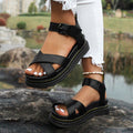 Sandales noires en cuir avec semelle épaisse, portées avec un jean blanc déchiré, posées sur un rocher près d'un étang. Mode estivale tendance.