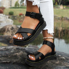 Sandales noires en cuir avec semelle épaisse, portées avec un jean blanc déchiré, posées sur un rocher près d'un étang. Mode estivale tendance.