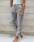 Pantalon en lin gris pour femme, taille élastique avec cordon, poches latérales, style décontracté, porté avec un haut blanc, fond en bois clair.