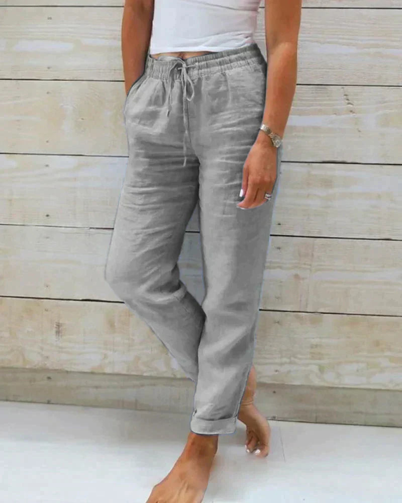 Pantalon en lin gris pour femme, taille élastique avec cordon, poches latérales, style décontracté, porté avec un haut blanc, fond en bois clair.