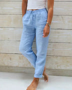 Pantalon en lin bleu clair pour femme, taille élastique avec cordon, style décontracté, porté avec un haut blanc, fond en bois clair.
