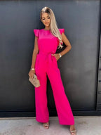 Femme en combinaison rose vif, posant devant un mur noir, tenant une pochette beige. Mode élégante, tenue chic, style moderne.
