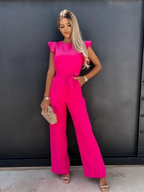 Femme en combinaison rose vif, posant devant un mur noir, tenant une pochette beige. Mode élégante, tenue chic, style moderne.