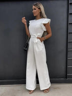 Femme portant une combinaison blanche élégante avec volants, sac à main noir, posant devant un mur sombre. Mode féminine chic et moderne.
