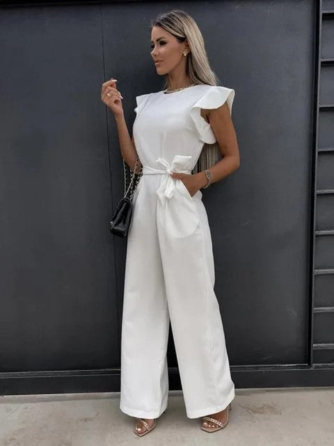 Femme portant une combinaison blanche élégante avec volants, sac à main noir, posant devant un mur sombre. Mode féminine chic et moderne.