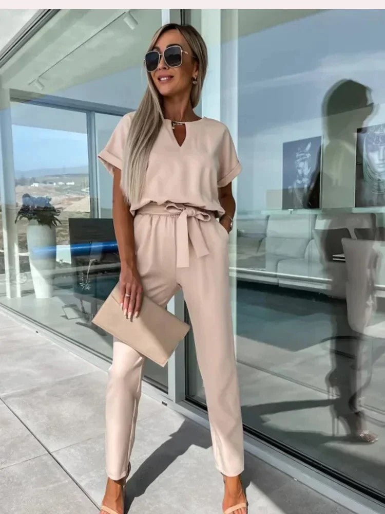 Femme élégante en combinaison beige, lunettes de soleil, tenant une pochette, debout sur une terrasse moderne avec vue panoramique. Mode chic et tendance.