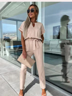 Femme élégante en combinaison beige, lunettes de soleil, tenant une pochette, debout sur une terrasse moderne avec vue panoramique. Mode chic et tendance.