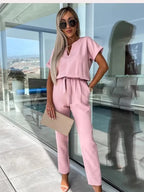 Femme en combinaison rose élégante, lunettes de soleil, tenant une pochette beige, debout devant une baie vitrée moderne. Mode féminine chic.