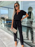 Femme élégante en tenue noire chic, lunettes de soleil, tenant une pochette beige, debout sur une terrasse moderne avec vue panoramique.