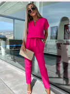 Femme en combinaison rose vif, lunettes de soleil, tenant une pochette beige, debout devant une baie vitrée moderne. Mode élégante et tendance.