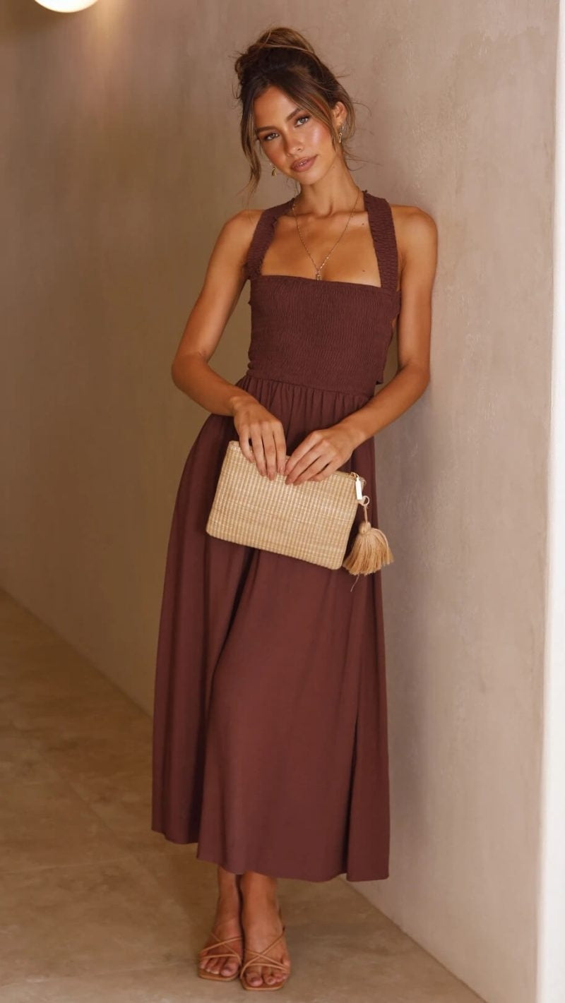 Femme en robe marron élégante, tenant une pochette beige, posant contre un mur beige. Mode féminine chic, tenue de soirée tendance.