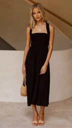 Femme en robe noire élégante, tenant un sac en osier, posant devant un mur beige. Mode estivale chic, style décontracté, tendance 2023.