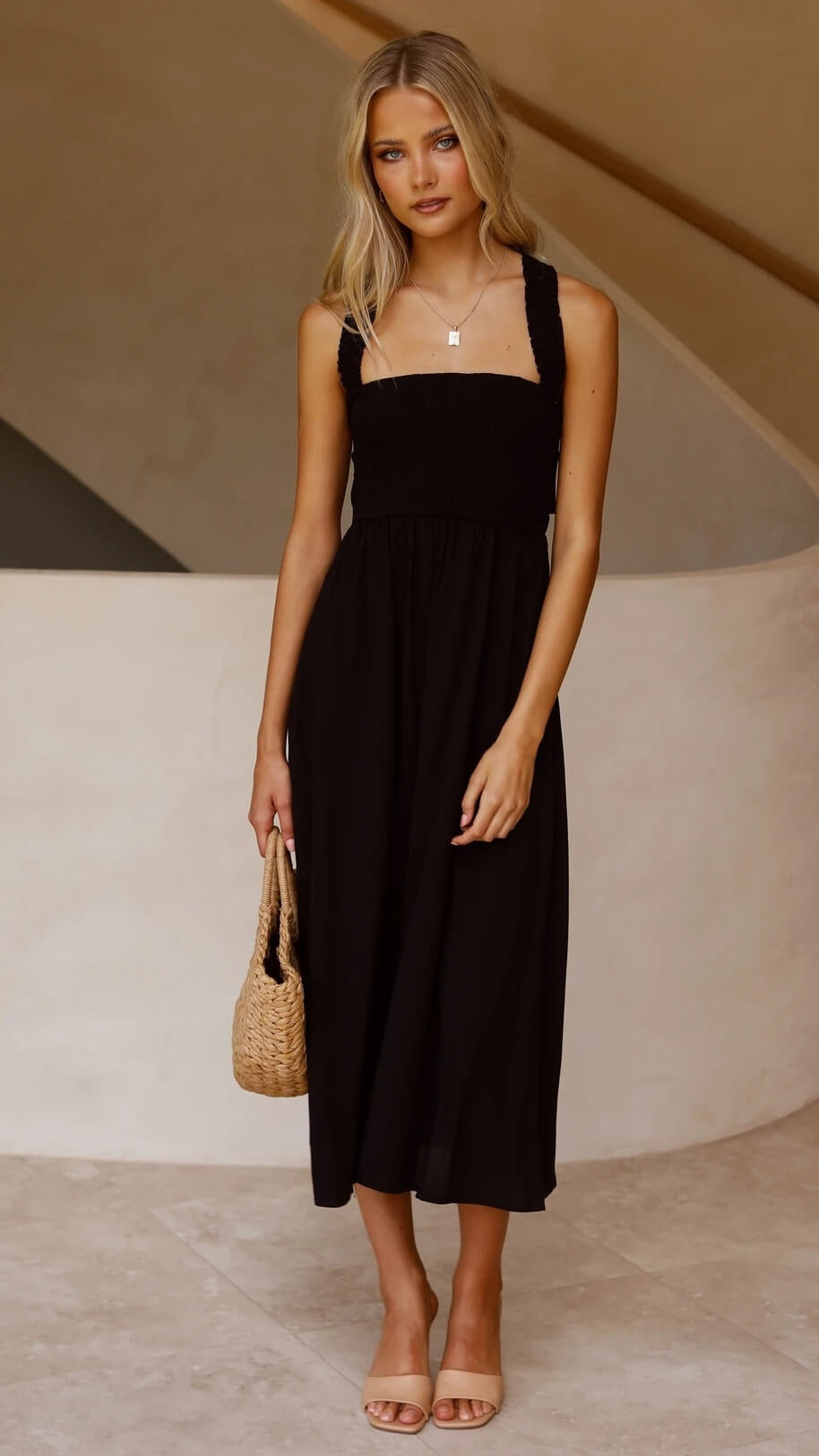 Femme en robe noire élégante, tenant un sac en osier, posant devant un mur beige. Mode estivale chic, style décontracté, tendance 2023.